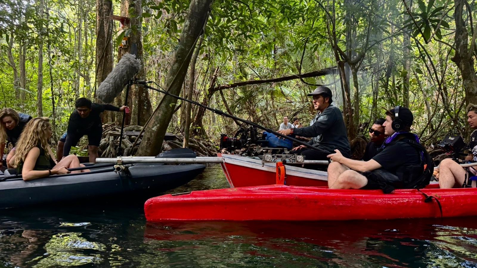 A sound recording setup in a jungle with two kayaks, featuring a sound technician using a boom microphone while working with a production crew. The scene includes individuals in both kayaks, engaged in a film shoot amidst lush greenery.
